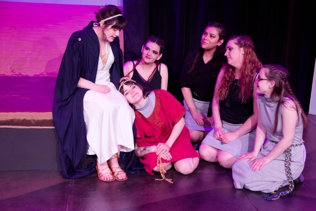 Ensemble in abstract Greek costumes: Penelope sits on the end of a bed, while an actress representing her son rests head on her knee. Four female servants listen.